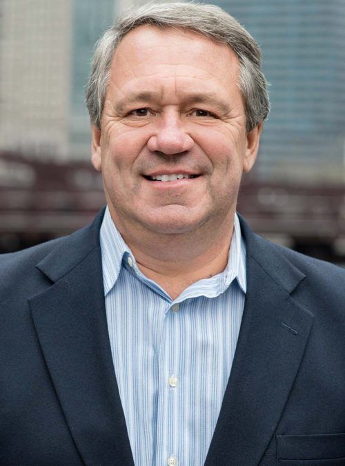 Smiling man in a navy blazer and blue striped shirt against a blurred urban background