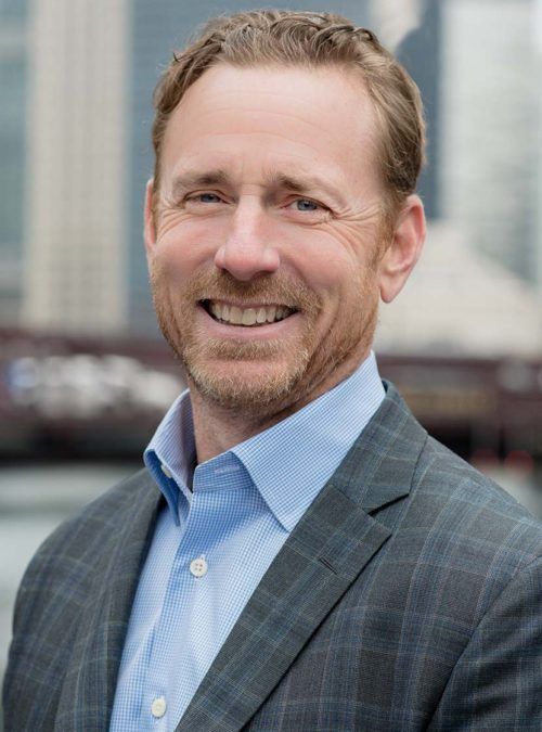 Smiling man in a blue shirt and gray blazer outdoors with blurred city buildings behind him