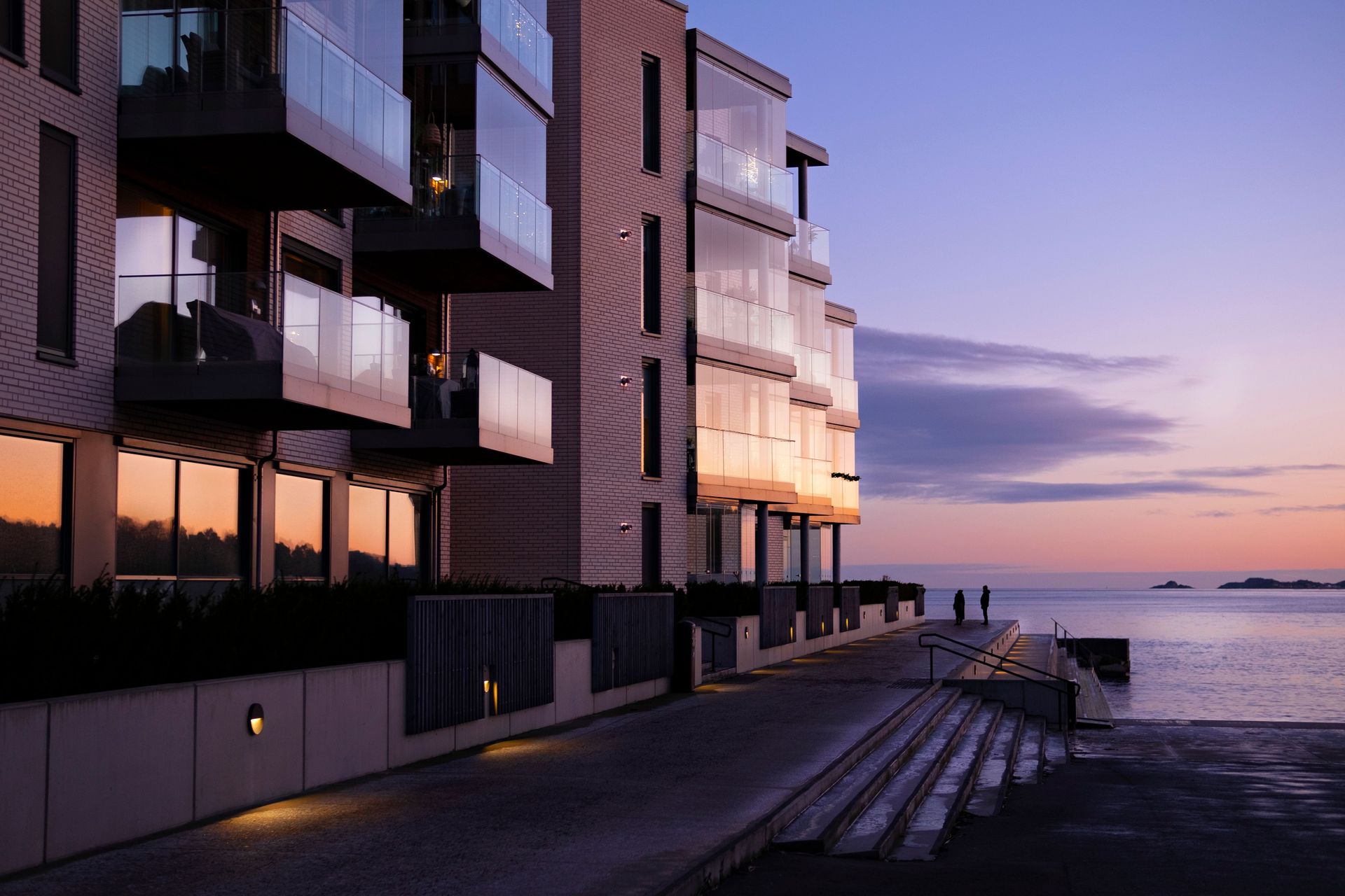 Modern waterfront apartment building at sunset beside a calm shoreline and walkway