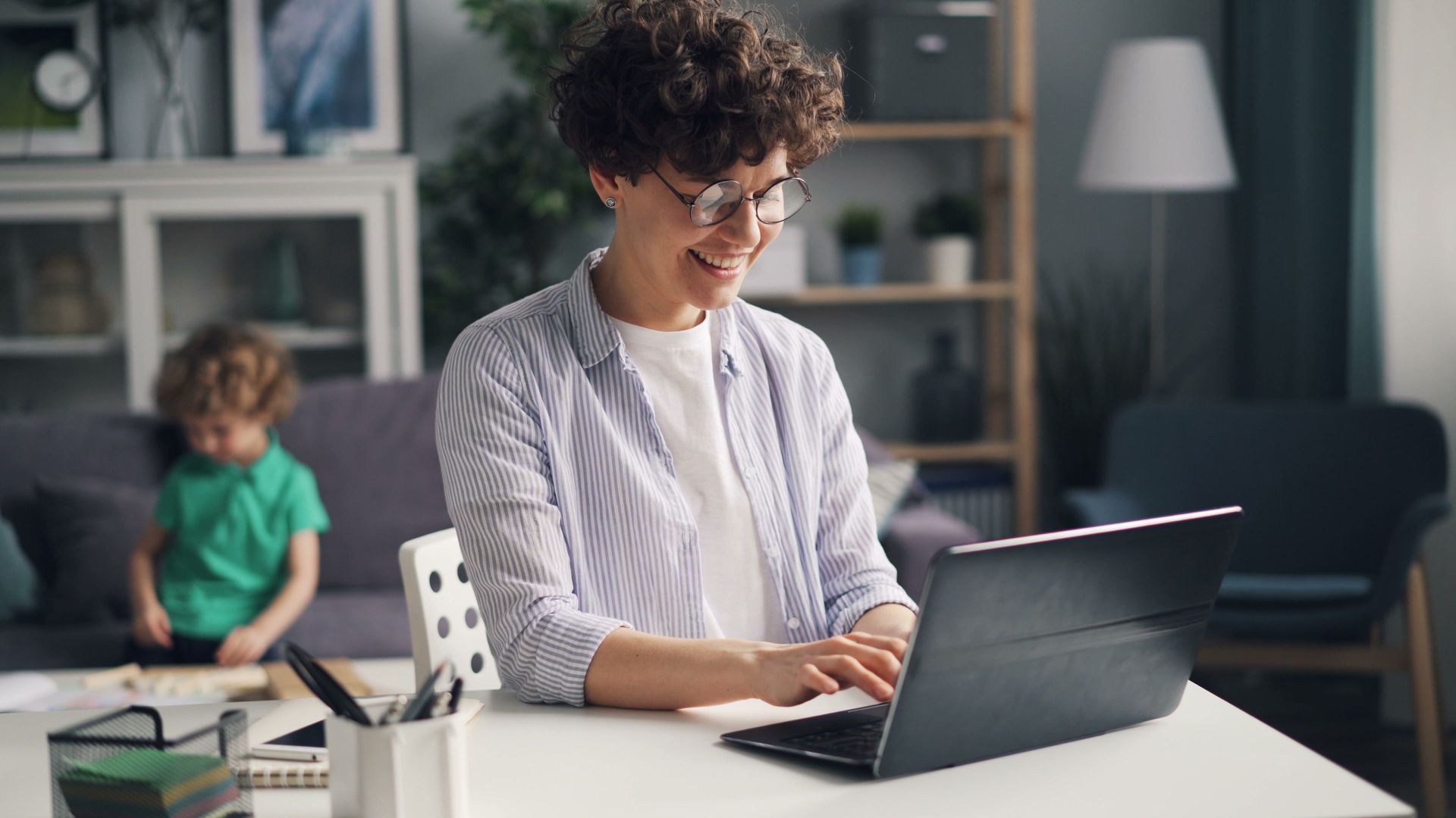 Person smiling and typing on a laptop at a white desk in a living room while a child sits in the background.