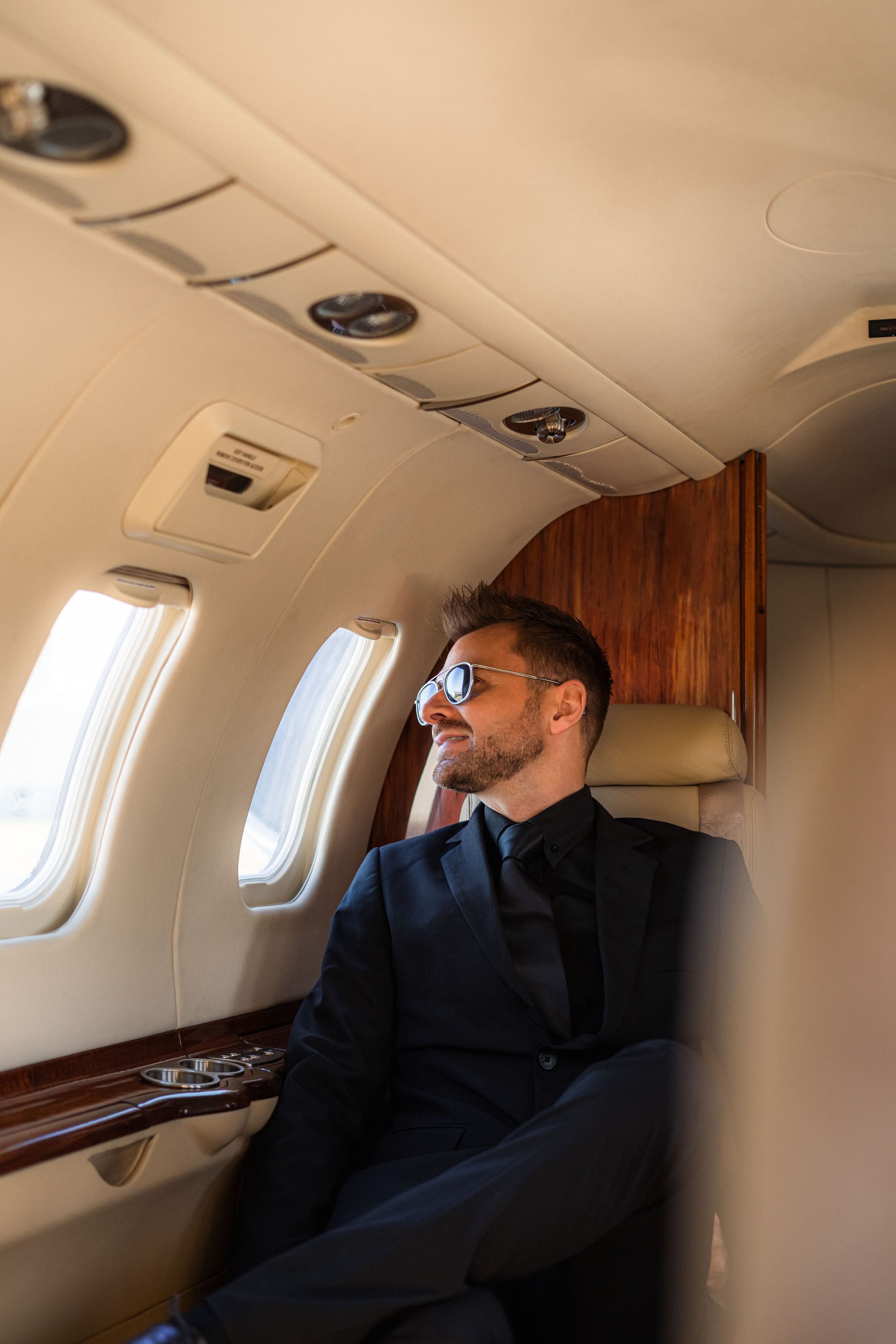 Man in dark suit and sunglasses seated on a private jet, looking out the window.