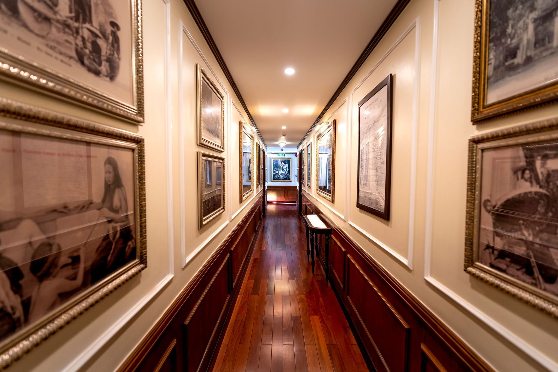 Narrow hallway with framed pictures on both walls and polished wooden floors, lit by ceiling lights