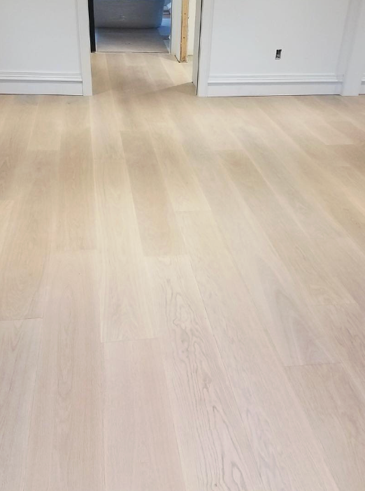 Light-colored, wide-plank hardwood flooring installed in a room with white baseboards and a doorway in the background.