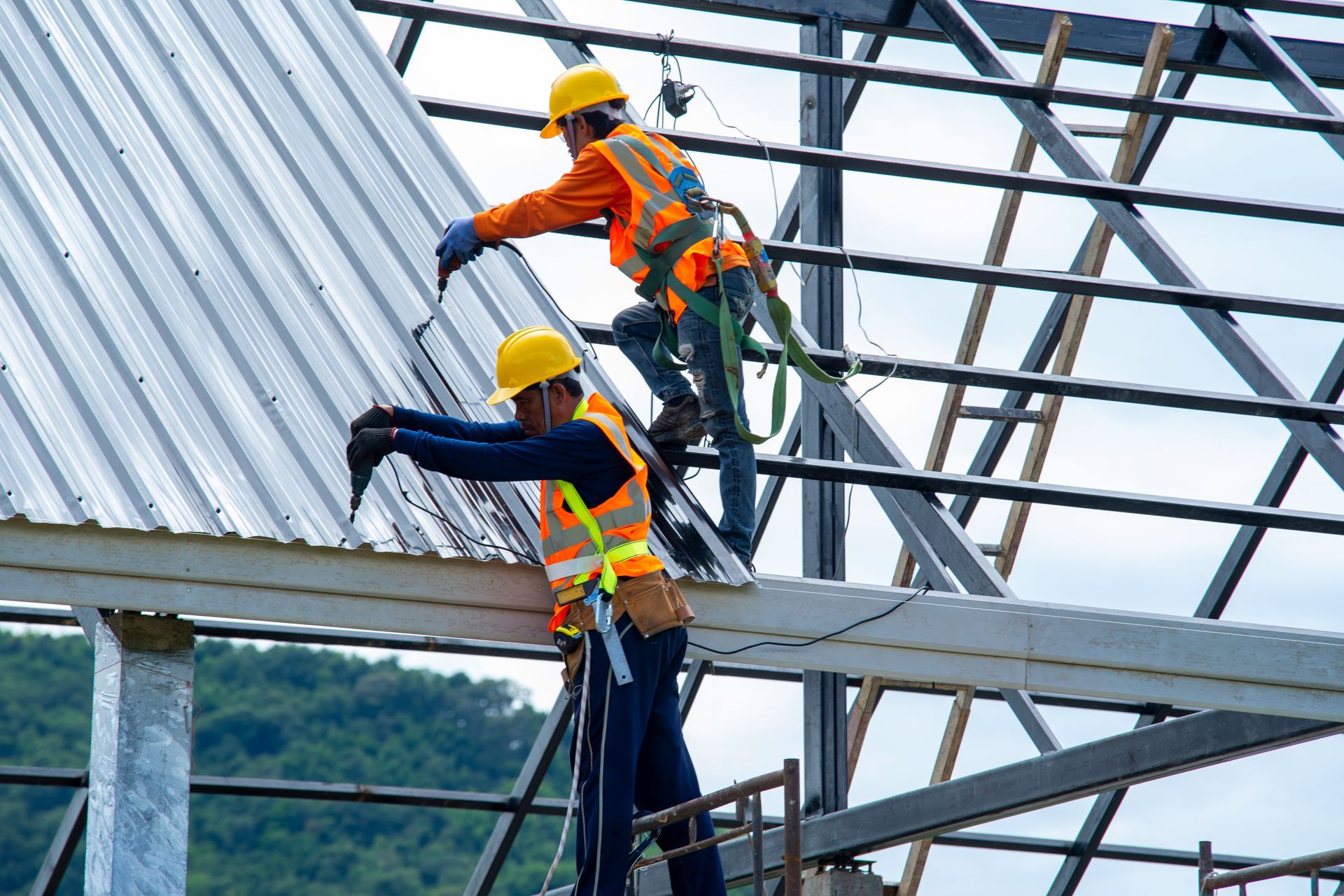 Two construction workers in safety gear installing a metal roof on a building frame.