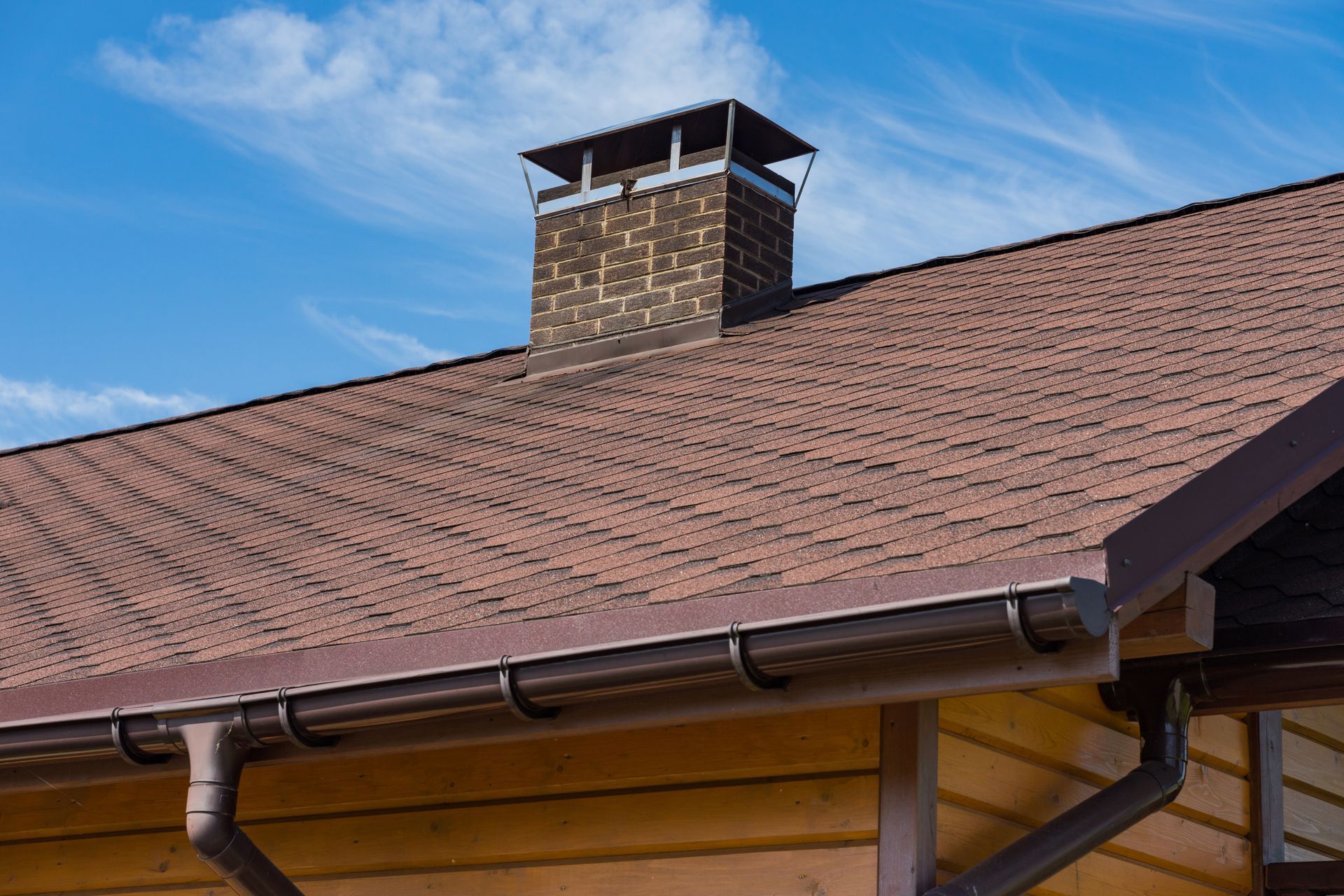 Brown shingle roof with chimney and gutters against a blue sky.