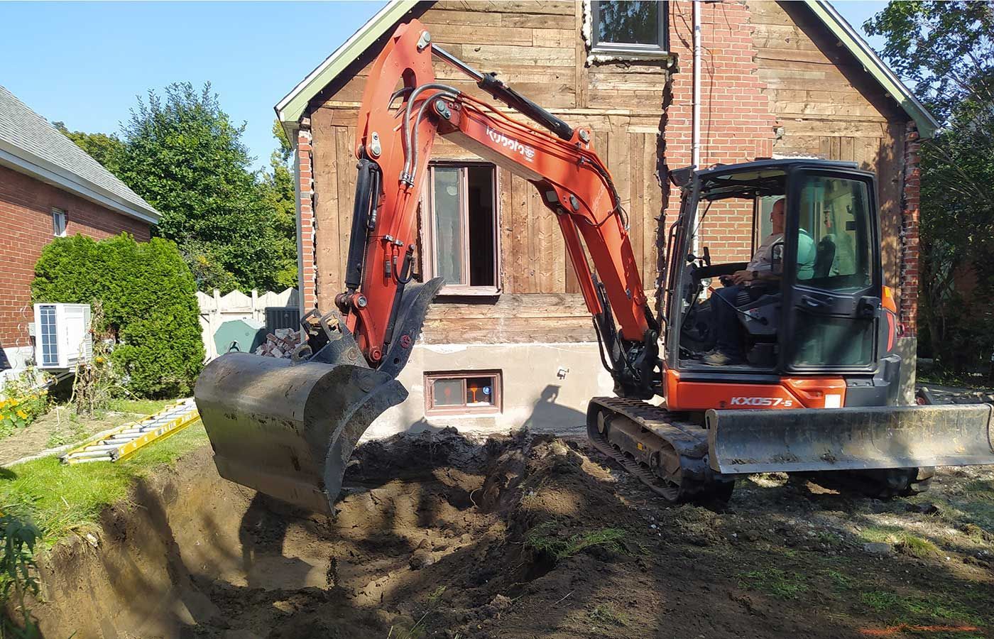 An orange excavator digs a trench in the yard in front of a house undergoing renovations.