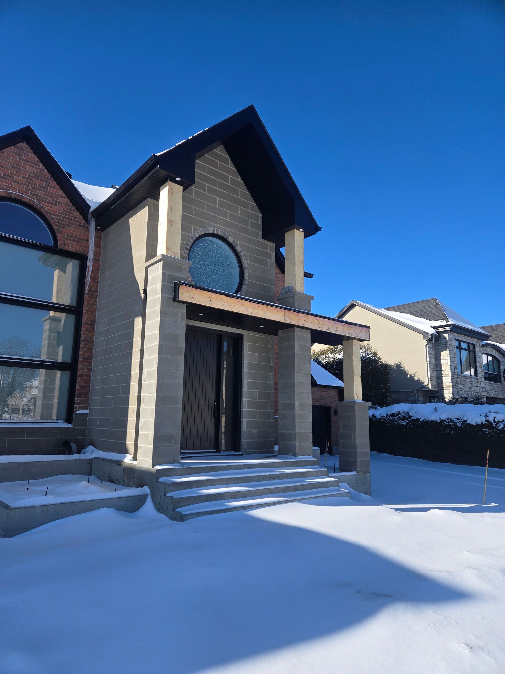 A modern house with a gray stone entrance, a circular window, and large glass panes, surrounded by deep winter snow.