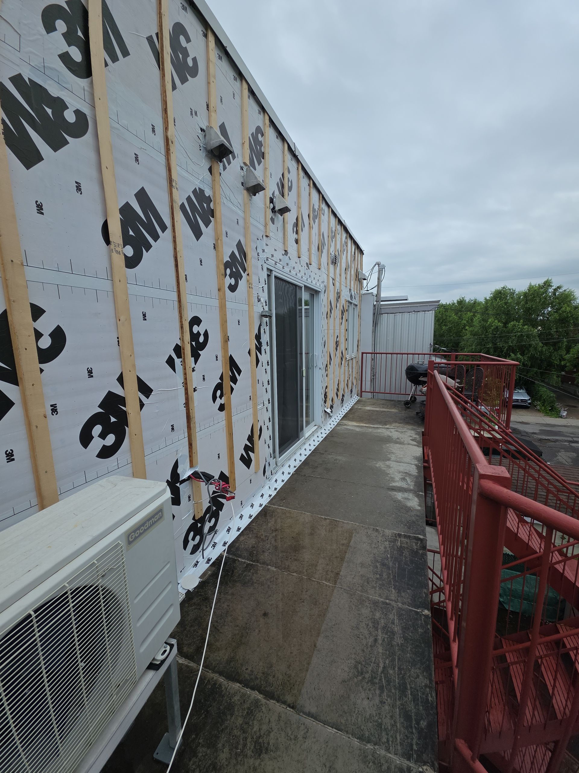 Exterior view of a building under renovation, showing 3M moisture barrier, wood furring strips, a doorway, and red railing.