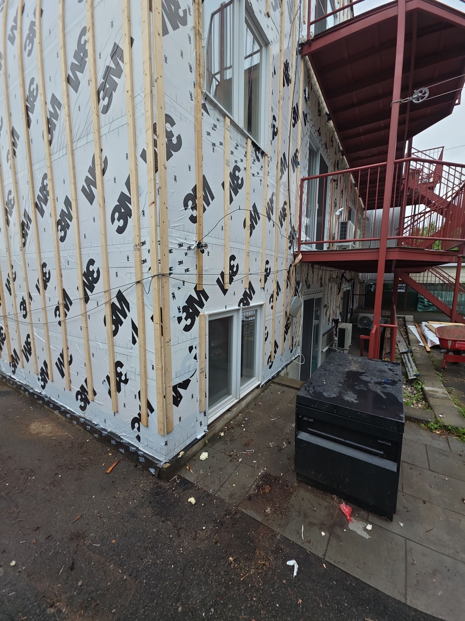Building exterior under construction with white protective wrap featuring 3M logos and vertical wood furring strips.