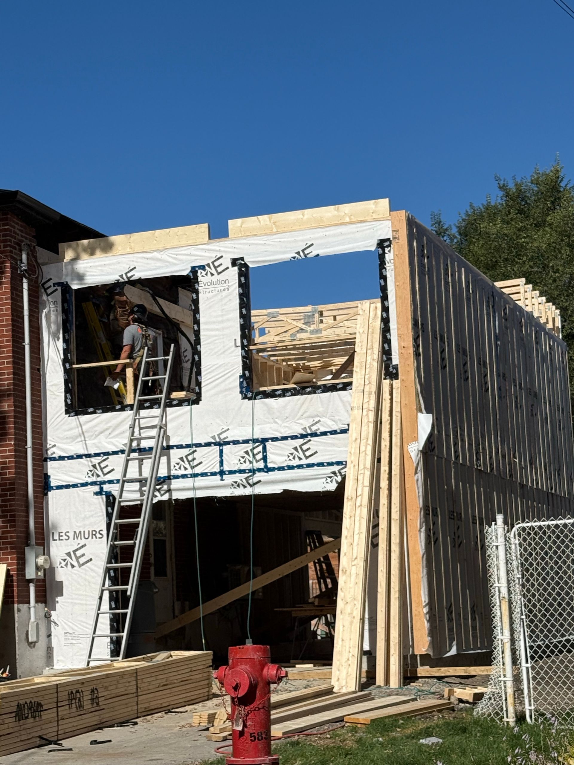 A person on a ladder working on the wooden framing of an unfinished two-story building under a clear blue sky.