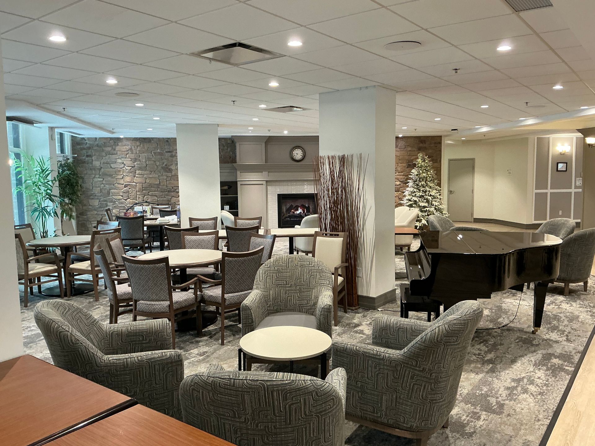 A well-lit hotel or residential lounge area with gray patterned armchairs, dining tables, a stone fireplace, and a piano.