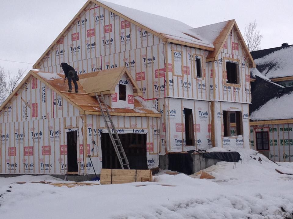 Un ouvrier se tient sur le toit d'une maison en construction, recouverte d'une bâche Tyvek, dans un décor enneigé.