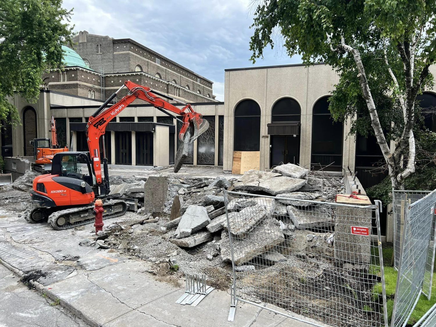 An orange excavator demolishes concrete rubble in front of a building with arched windows.
