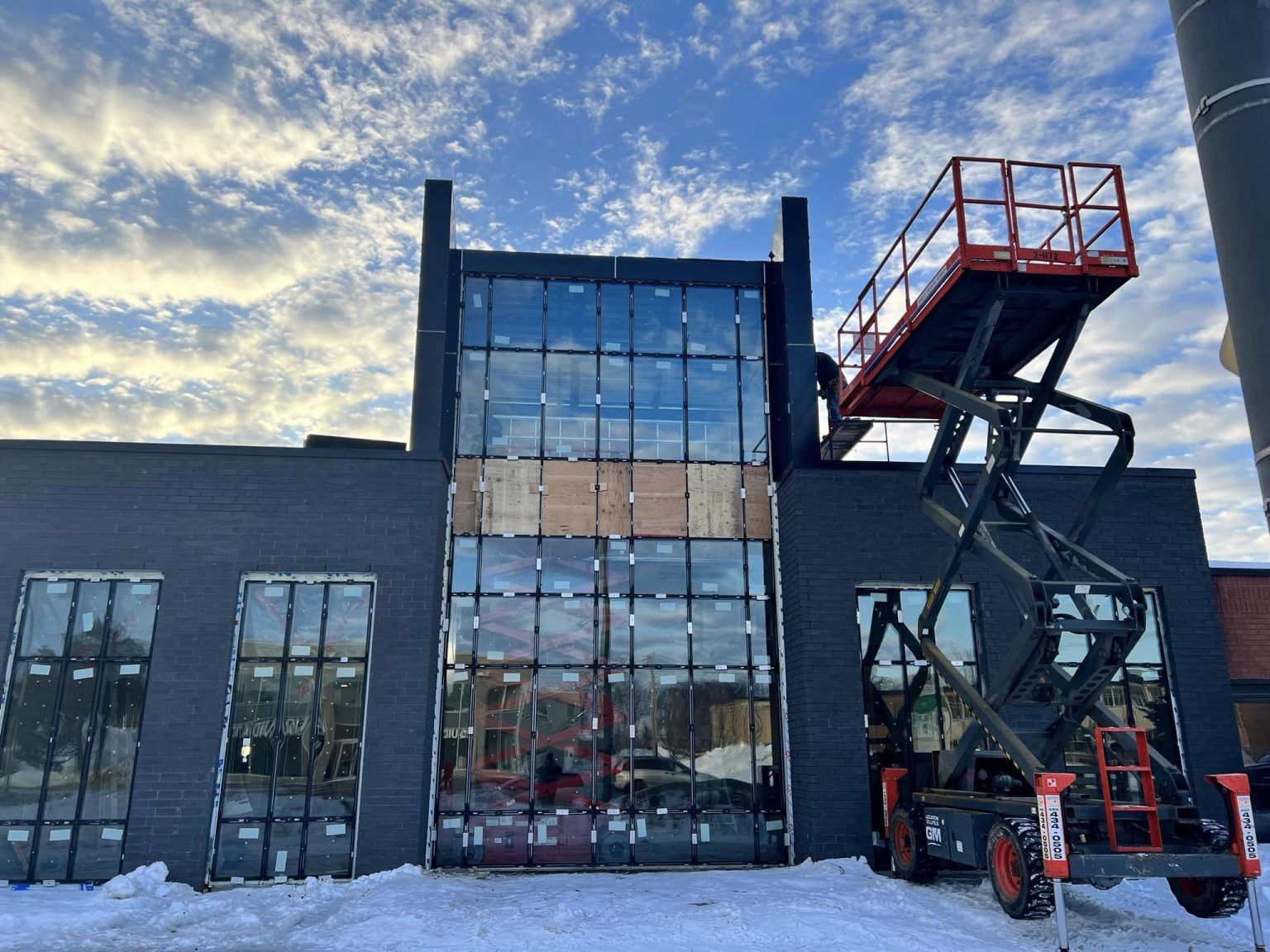 A scissor lift sits parked in front of a dark building with large window panels under a partly cloudy sky.