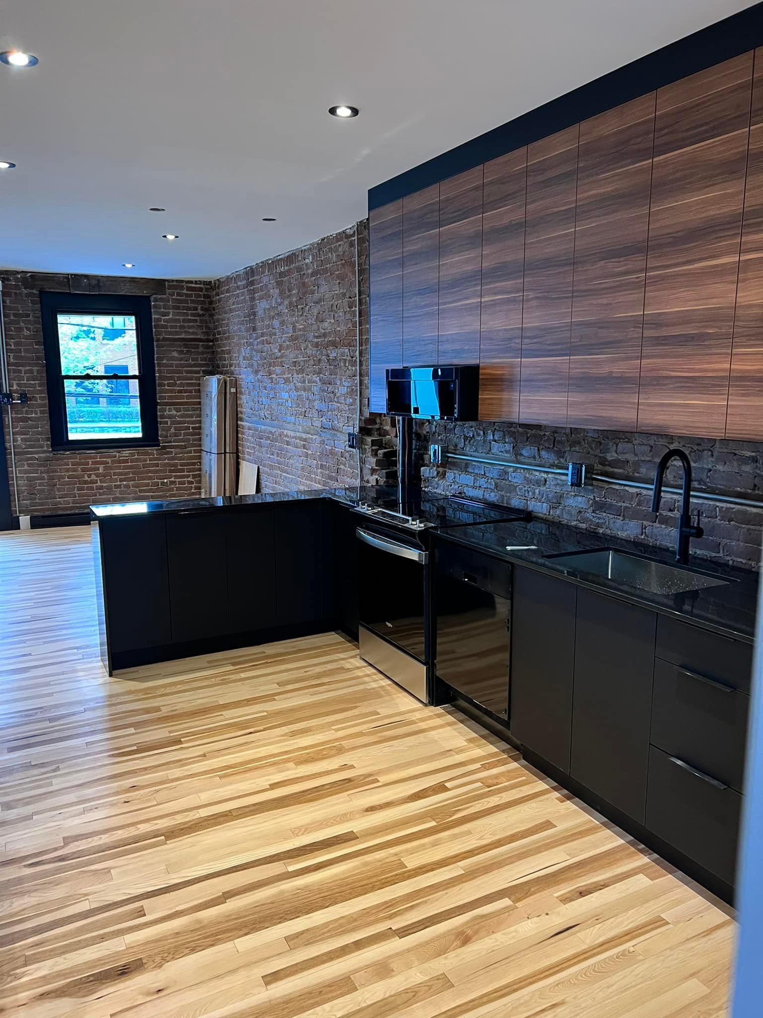 A modern kitchen featuring black cabinets, a dark wood-paneled backsplash, an exposed brick wall, and light wood flooring.