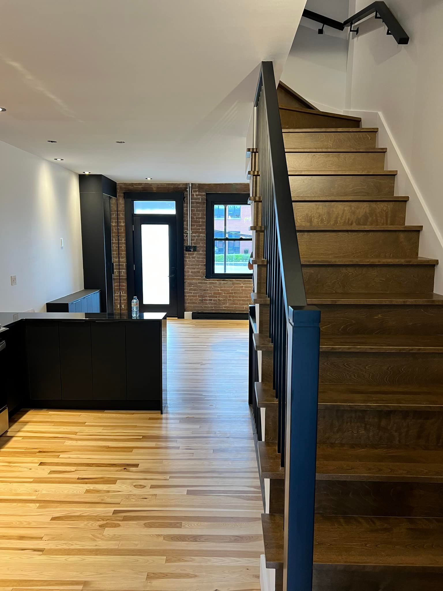 A modern, open-concept living space with light wood floors, black cabinetry, a brick accent wall, and a wooden staircase.