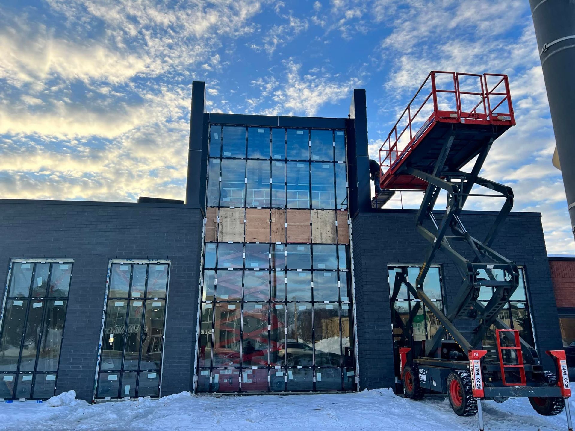 A black brick building under construction with glass panels and a red scissor lift parked in front against a cloudy sky.