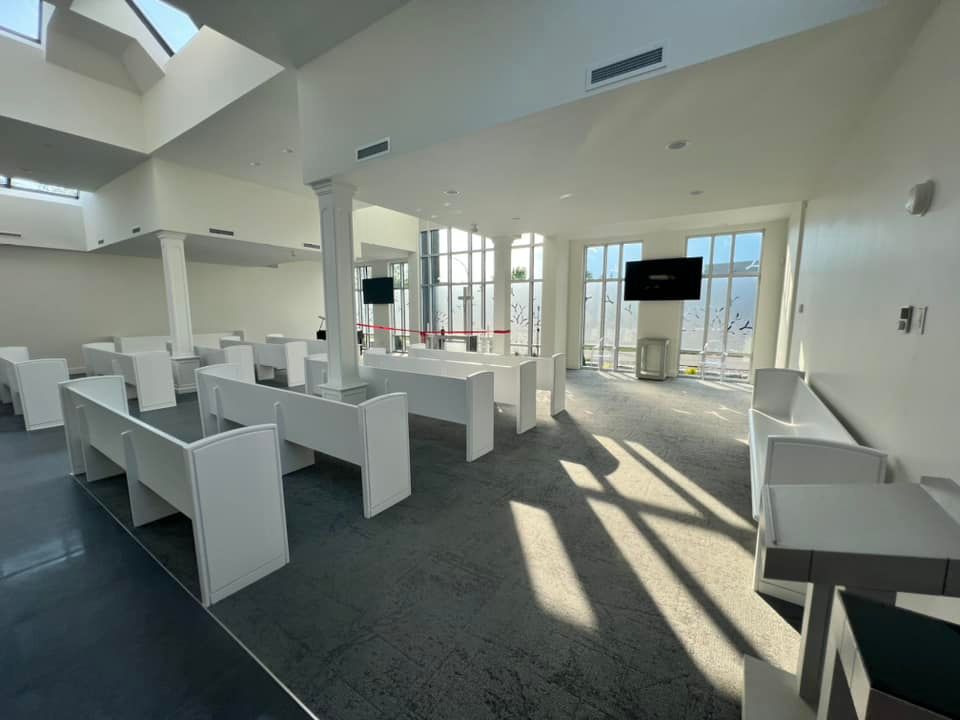A bright, modern event space with rows of white benches, light gray carpet, and large windows with natural sunlight.