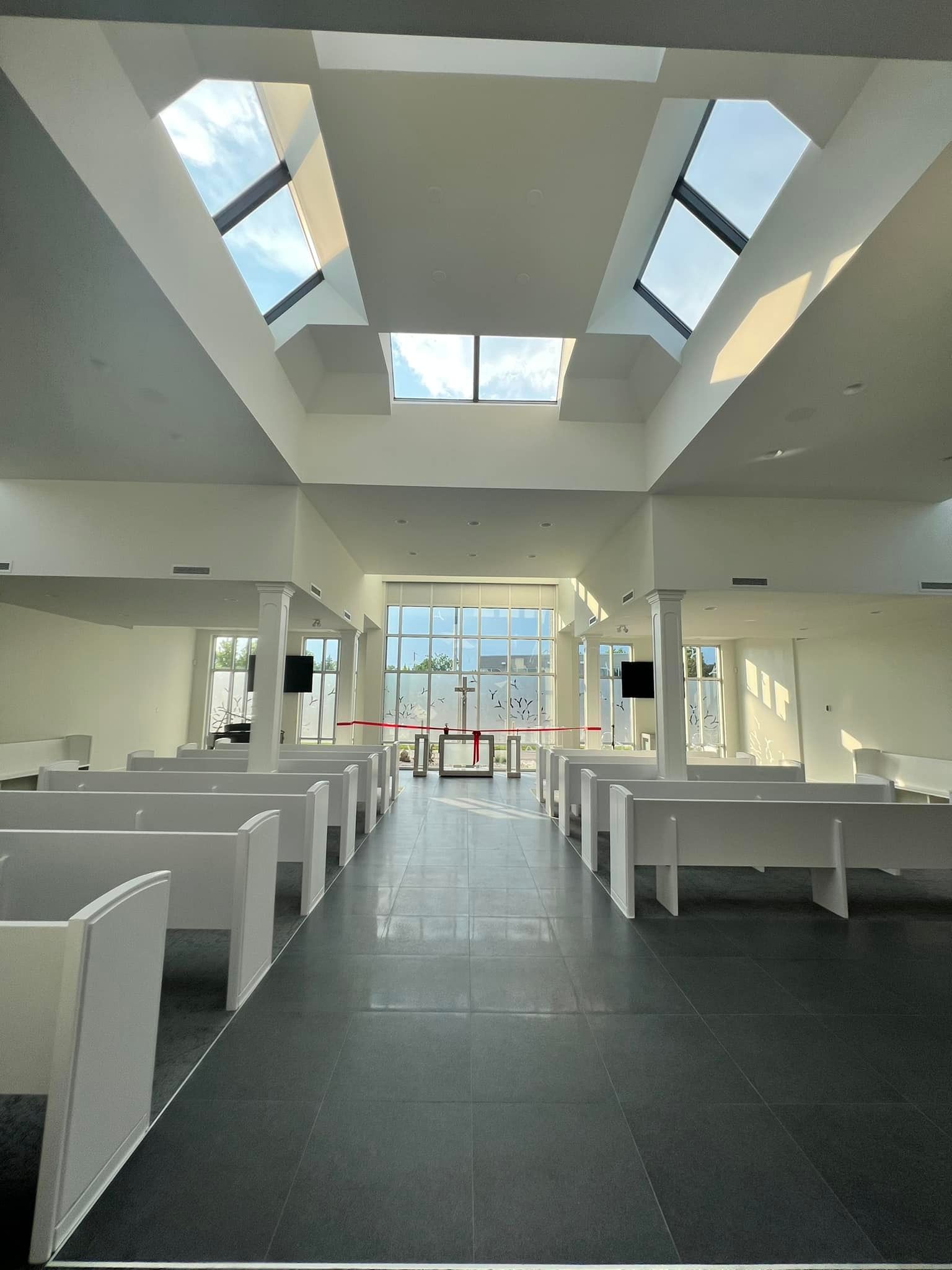 An interior view of a modern chapel with rows of white pews, dark tile flooring, and a ceiling with bright skylights.