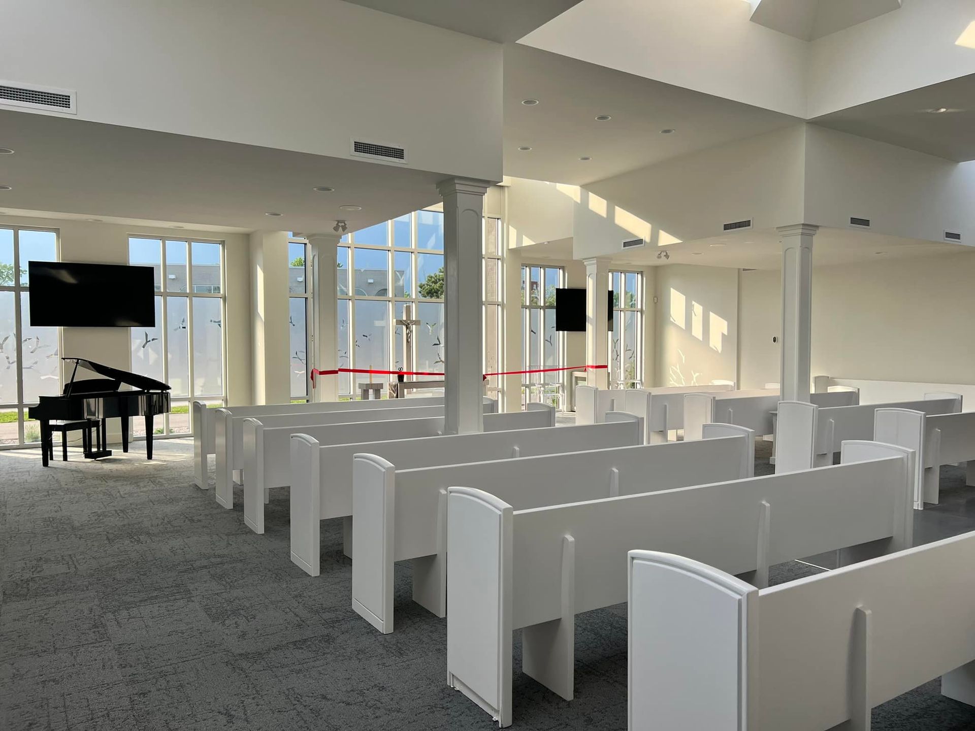 A bright, modern interior of a chapel with white pews facing a grand piano and a television mounted on the wall.
