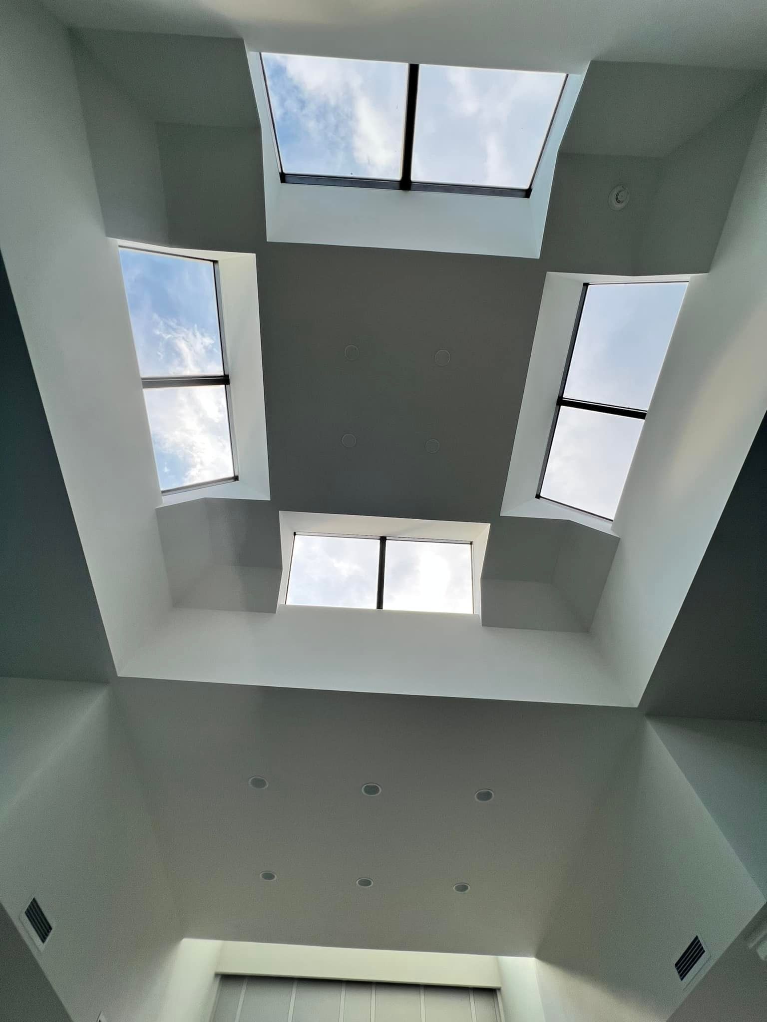 A modern, white-walled atrium featuring four square skylights looking up at a bright, cloudy sky.