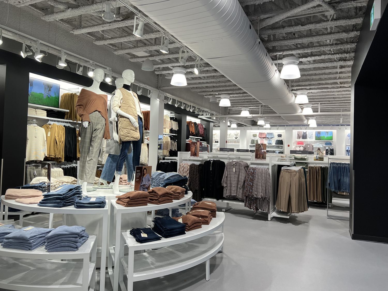 A clothing store interior featuring displays of folded apparel, racks of hanging garments, and mannequins.