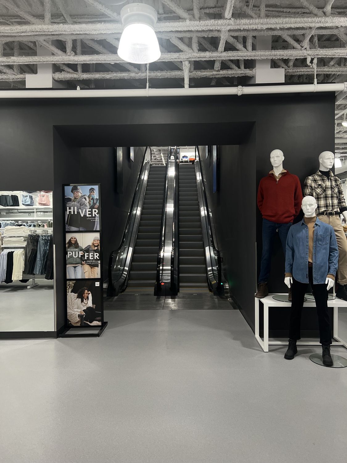 An indoor store entrance featuring two escalators framed by a black wall, a clothing display rack, and three mannequins.