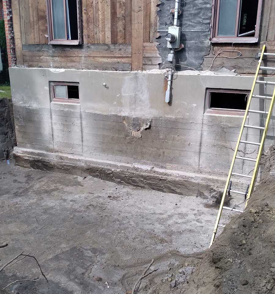 A side view of a house foundation with a basement excavation, showing gray concrete walls and a leaning yellow ladder.