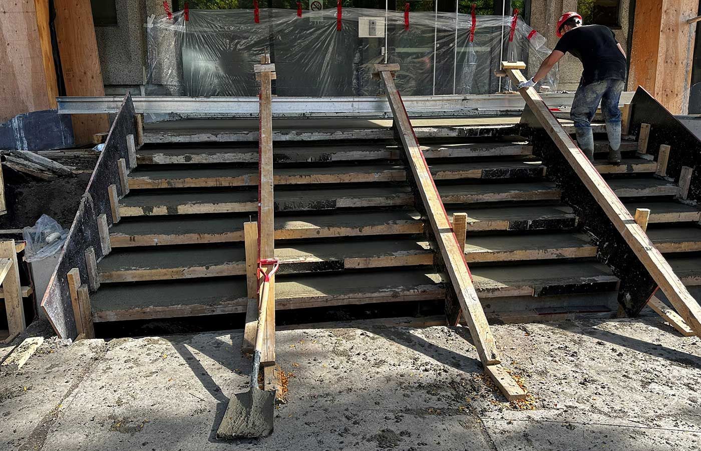 Construction workers pour concrete into wooden forms for a new staircase at a building entrance.