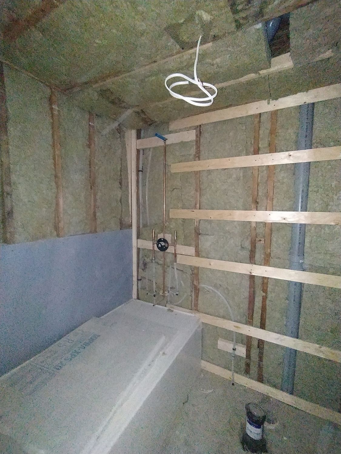 A bathroom under construction showing a bathtub, exposed wooden wall studs with insulation, and a shower valve installed.