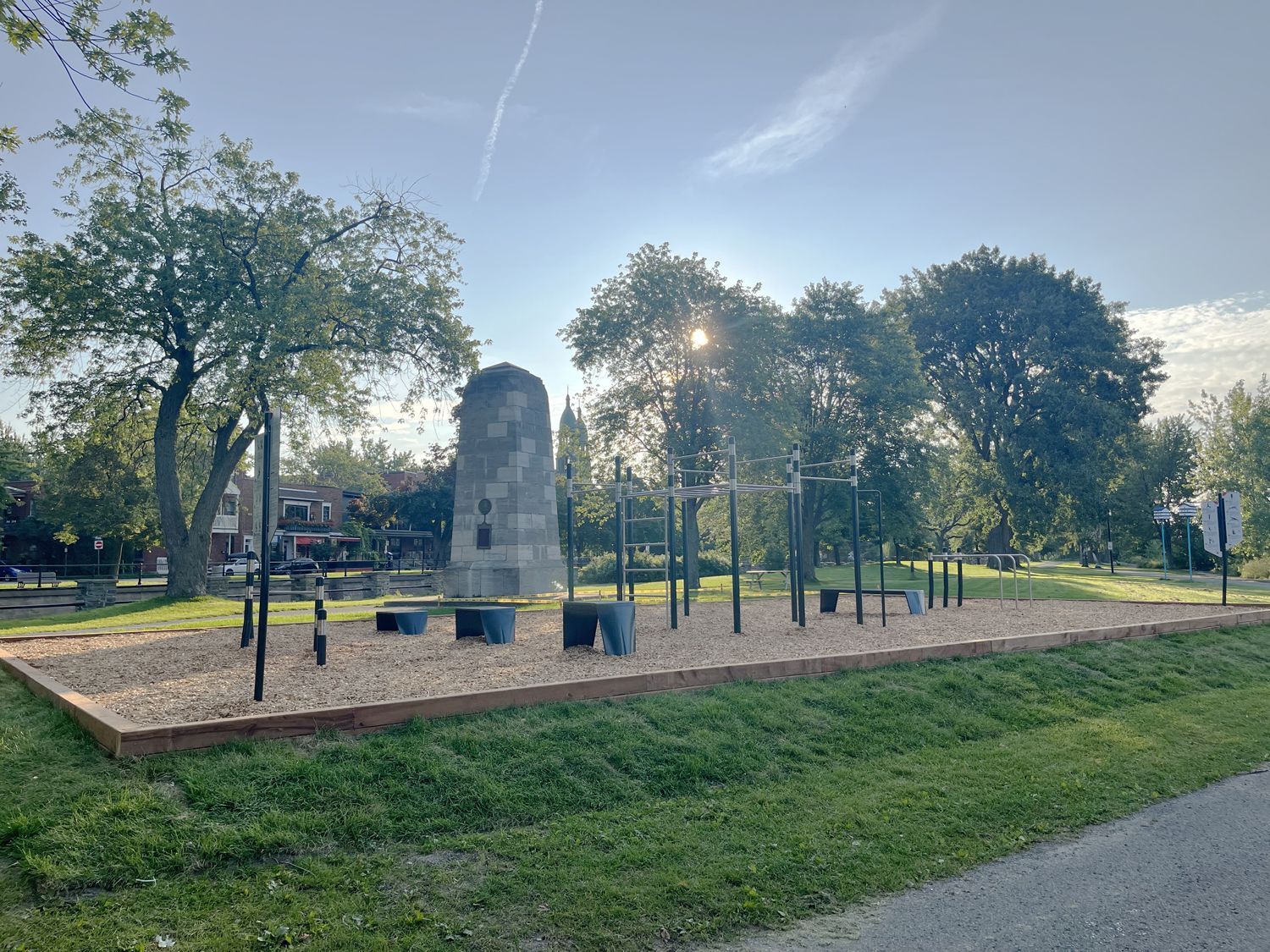 An outdoor calisthenics gym with black exercise bars on wood chips, featuring a stone monument in the background.