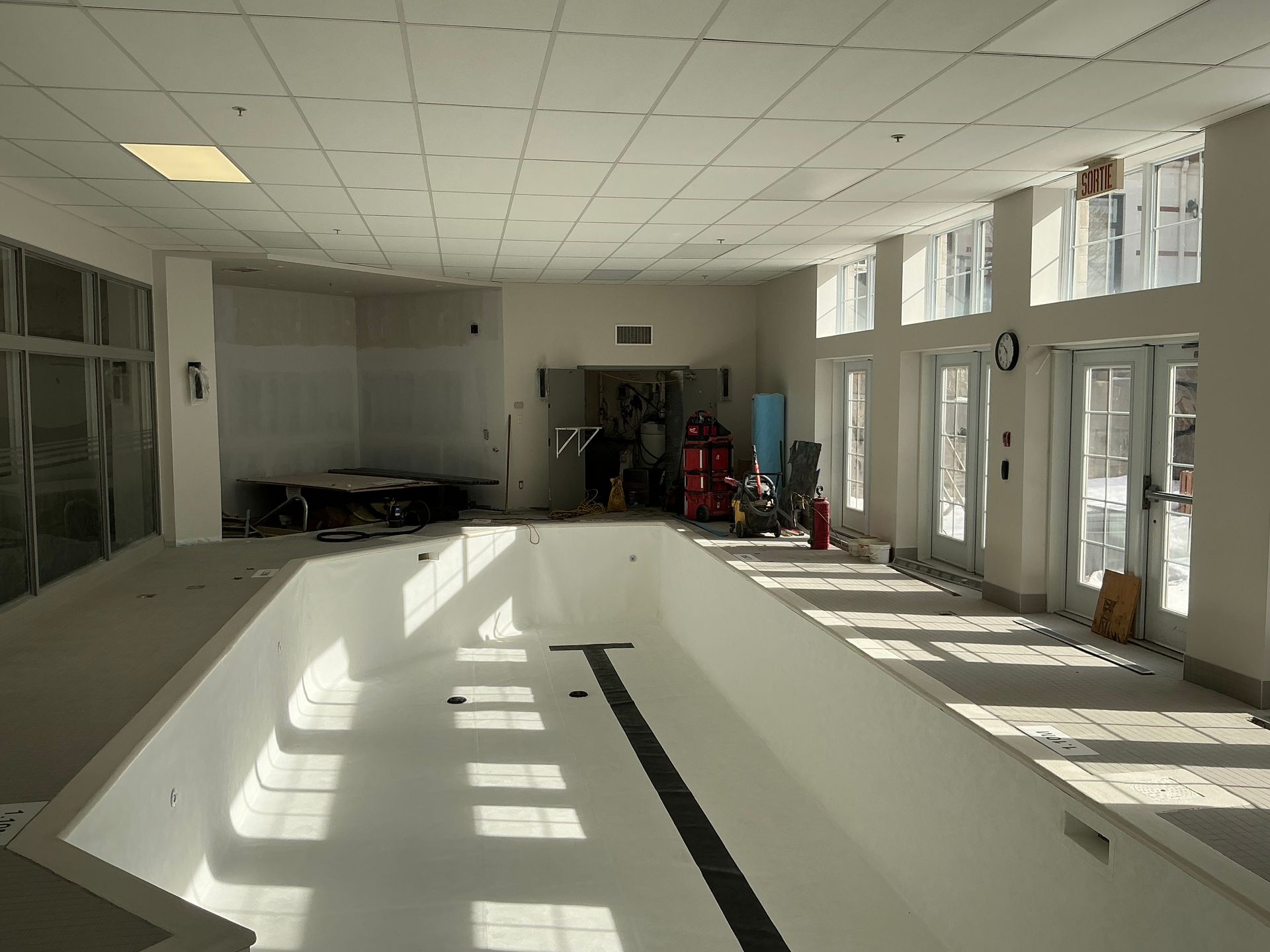 An indoor pool area under renovation, featuring a white empty pool with a black lane marker and large bright windows.