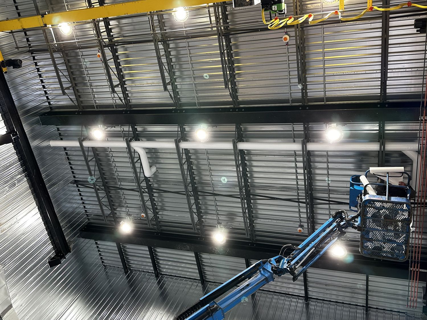 A blue aerial work platform lift reaches up toward an industrial ceiling with metal beams, piping, and lighting fixtures.