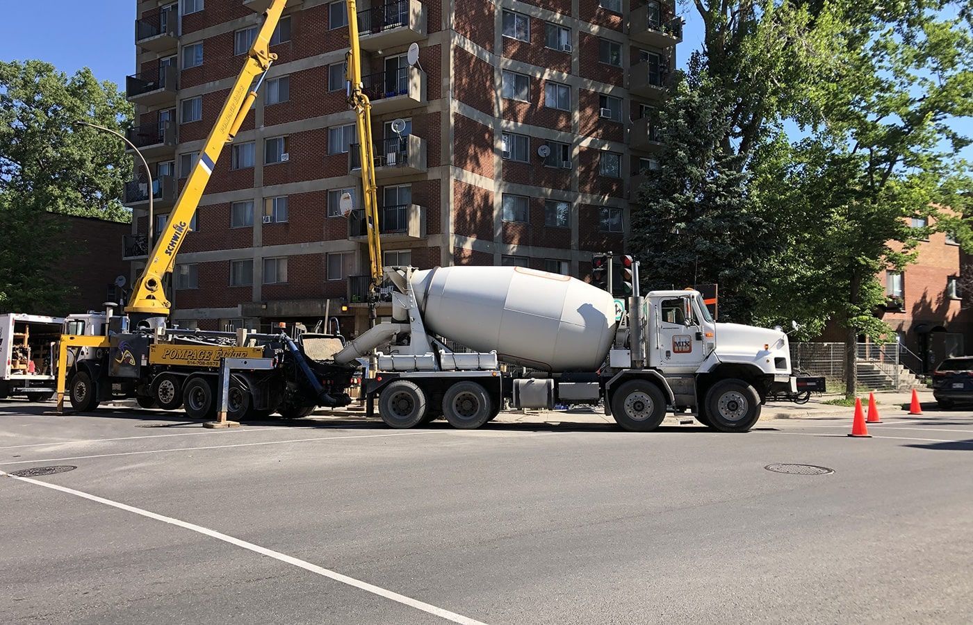 A white concrete mixer truck and a yellow crane parked on a street in front of a multi-story apartment building.