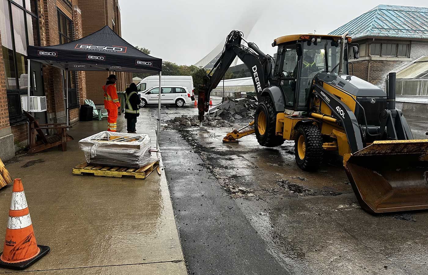 A yellow backhoe loader sits on an outdoor job site near a black canopy tent, a traffic cone, and construction workers.