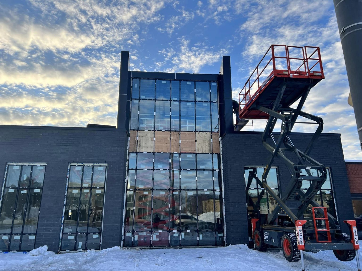 Un bâtiment sombre et moderne en construction, avec des fenêtres en verre et une nacelle élévatrice rouge garée devant, se détachant sur un ciel enneigé.