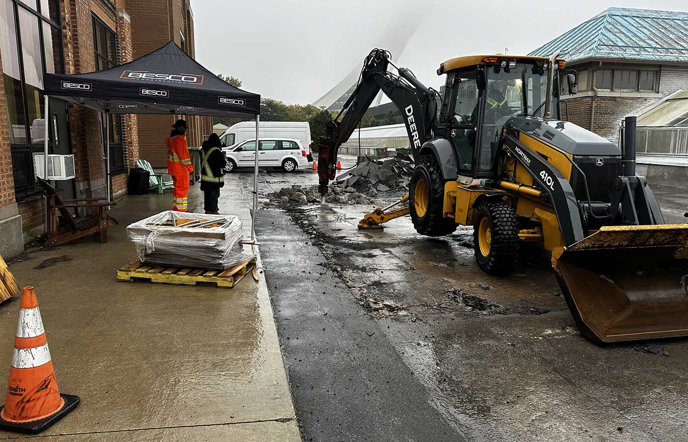Une pelleteuse de chantier jaune est en activité près de l'entrée d'un bâtiment, des personnes se trouvant à proximité sous une tente et un cône de signalisation orange.