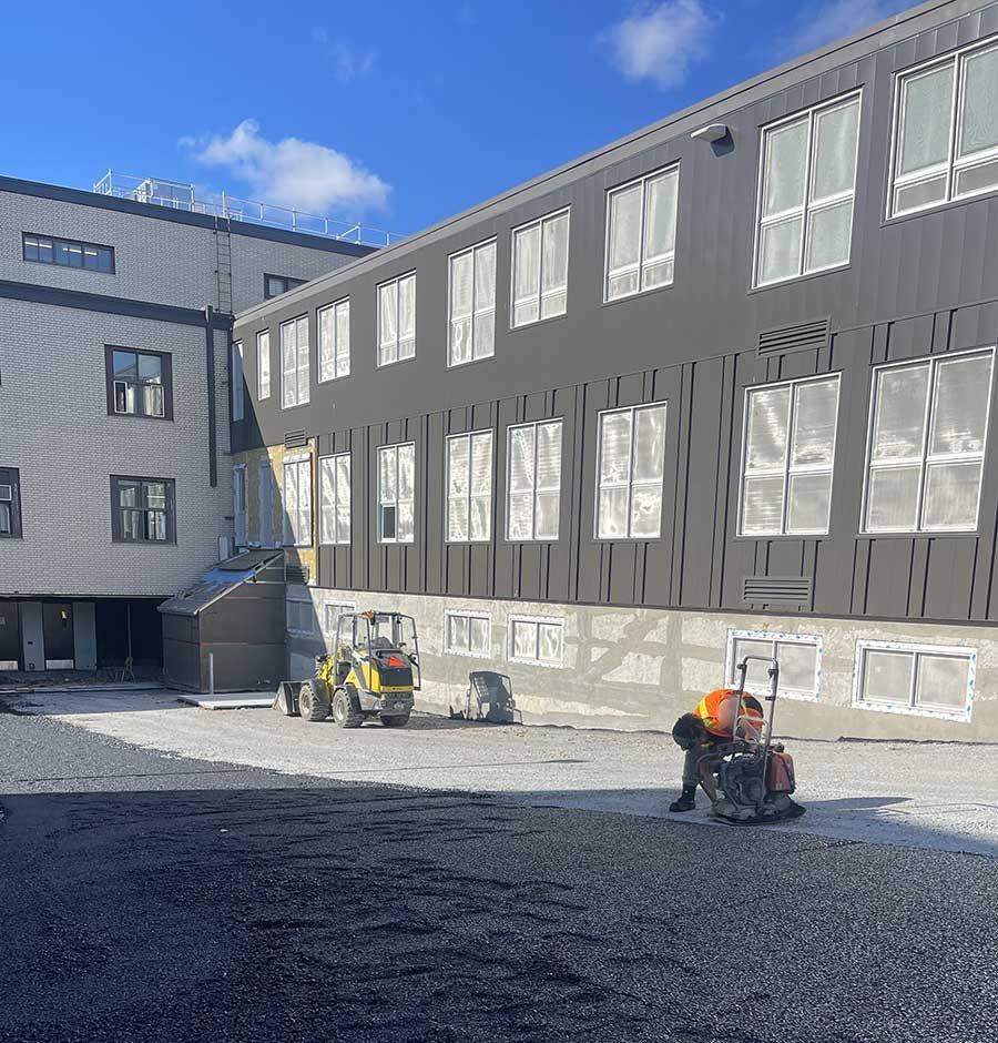 A construction worker uses a plate compactor on a gravel lot near a building with dark gray and light stone siding.