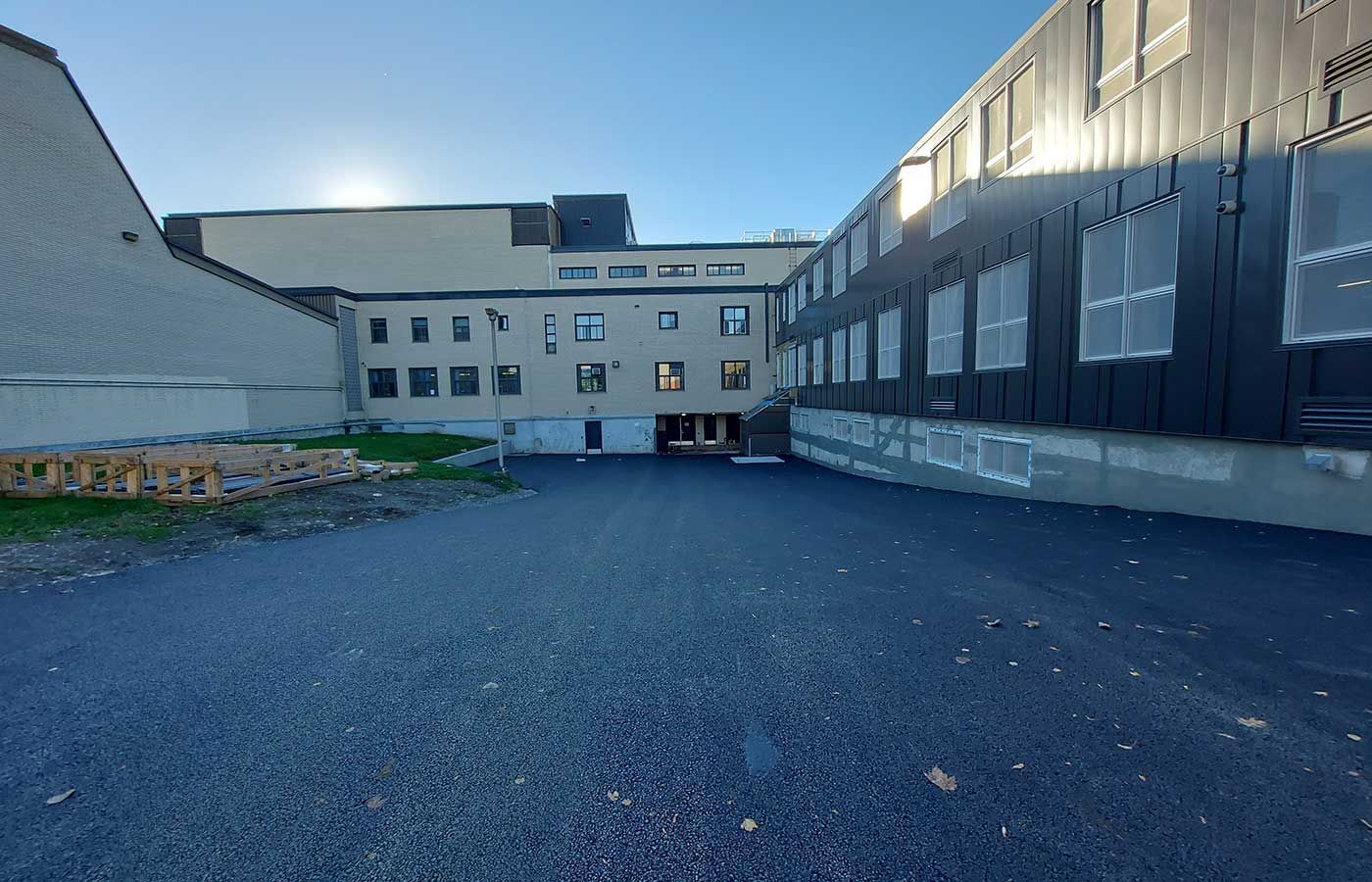 A paved courtyard between a light-colored building and a black building under a bright sky.