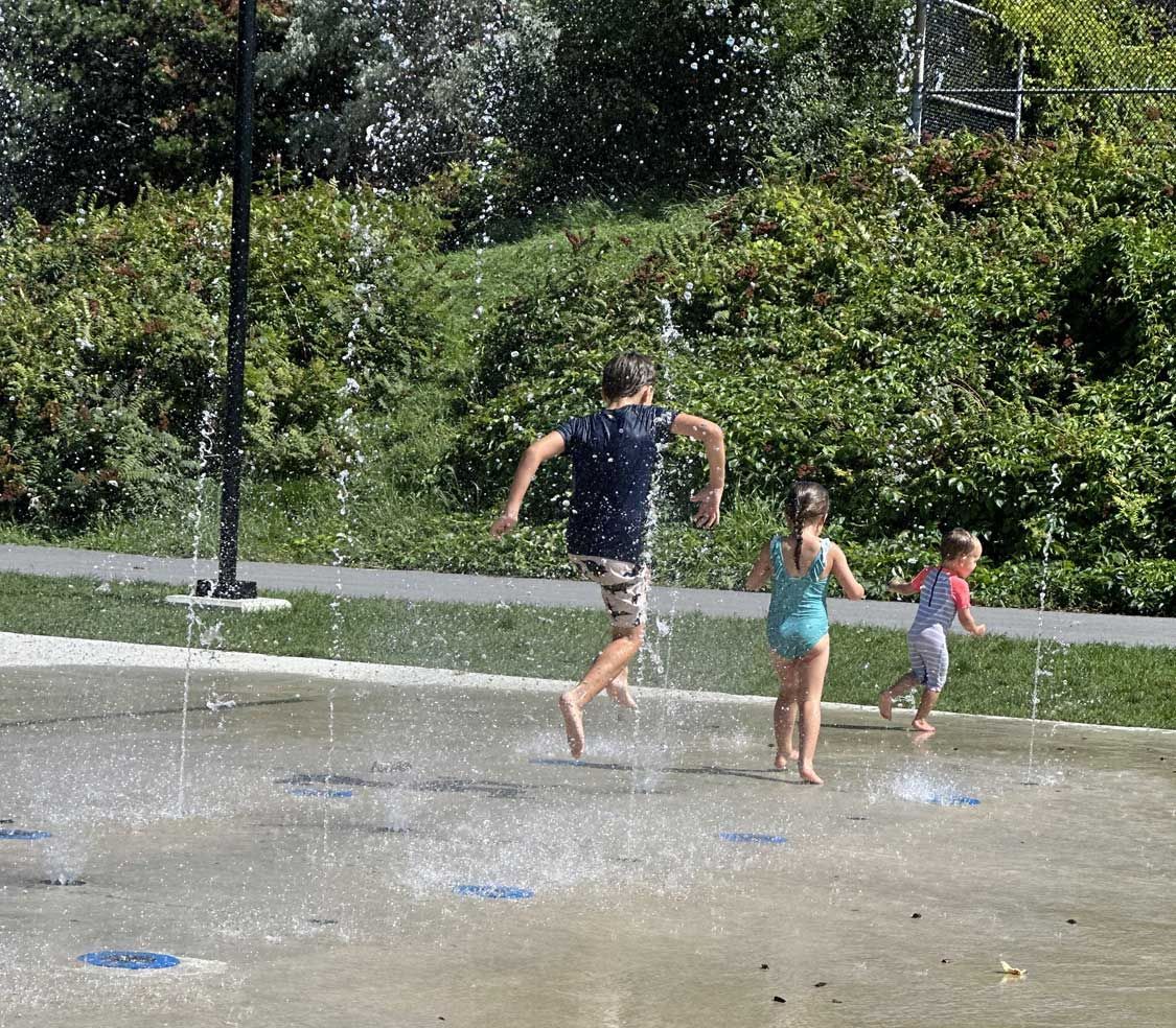Three children playing in a park splash pad with water jets spraying upwards, surrounded by lush green trees.