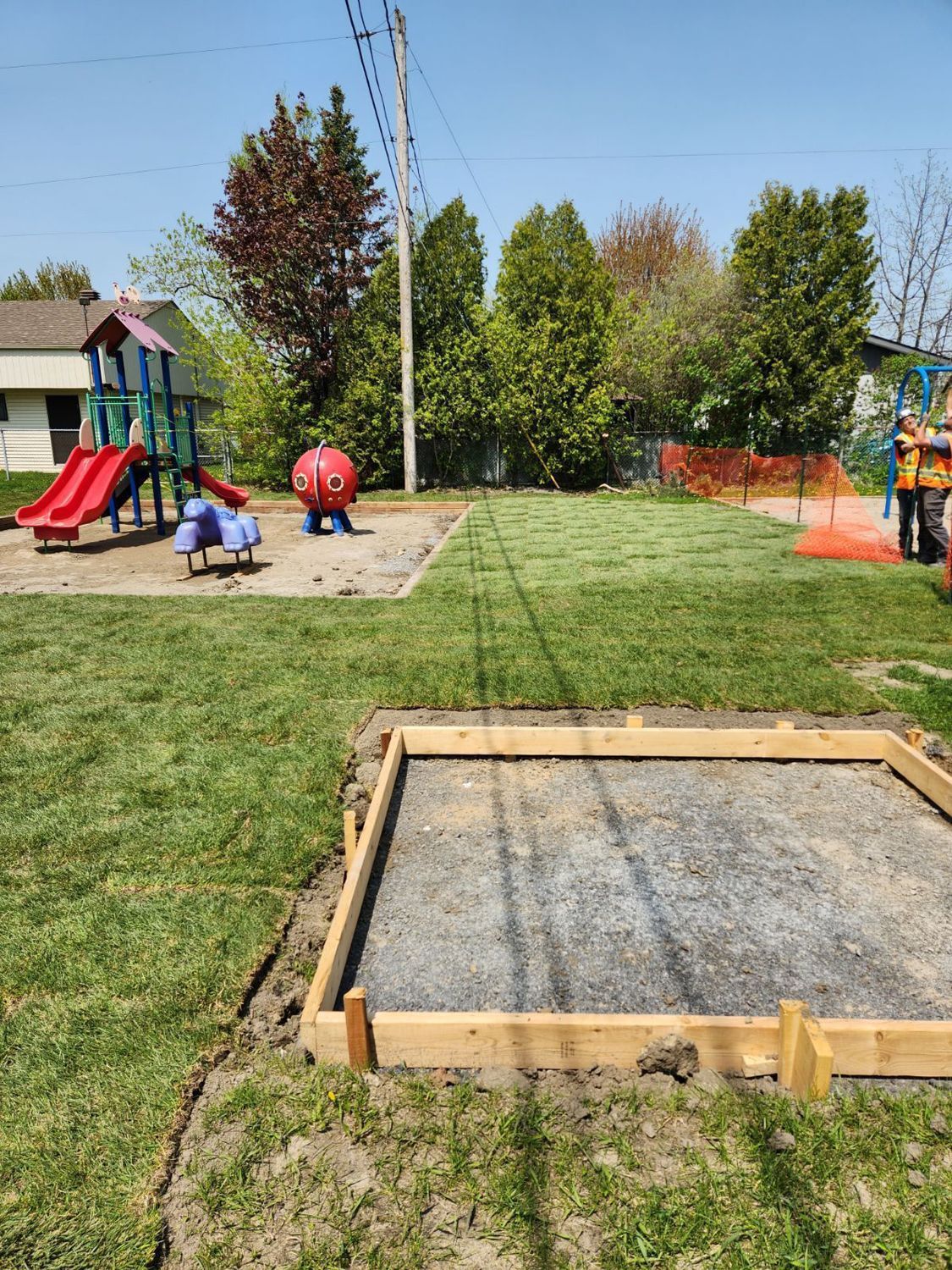 A playground under construction with a wooden frame around a gravel square, a slide set, and new sod on a sunny day.
