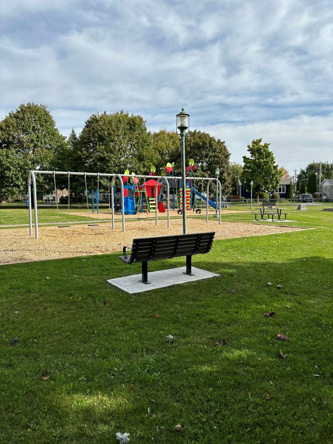 A black metal bench sits on a concrete pad facing a wooden-chipped playground with swings and a slide under a sunny sky.