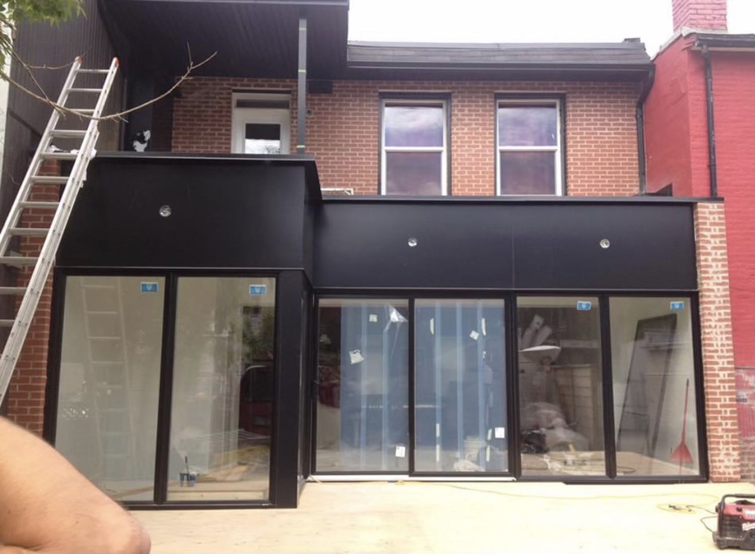 Back of a house with a modern black addition, large glass doors, a brick upper level, and a ladder leaning on the side.
