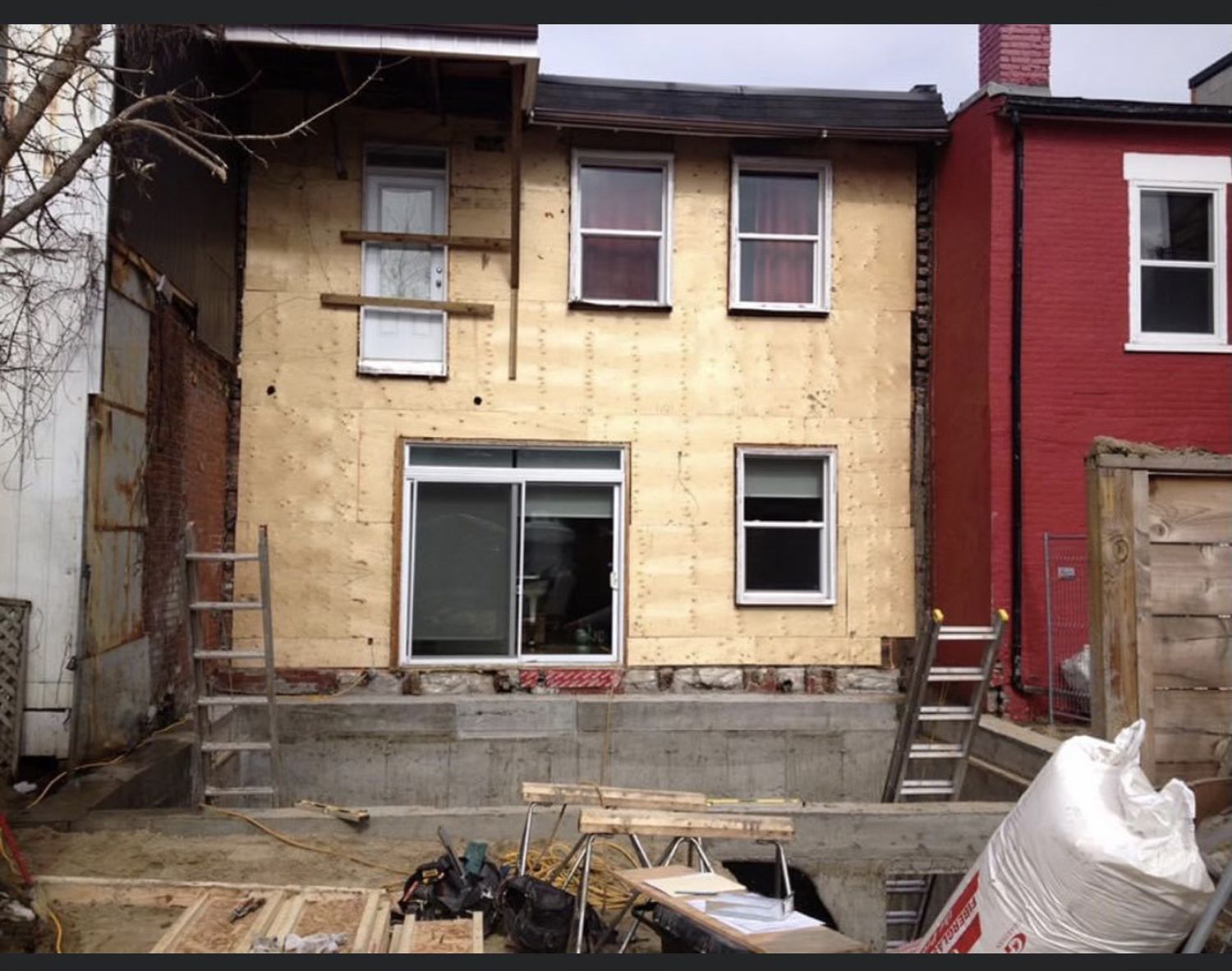 A residential home under construction, featuring exposed plywood walls, new windows, and a concrete foundation in a yard.