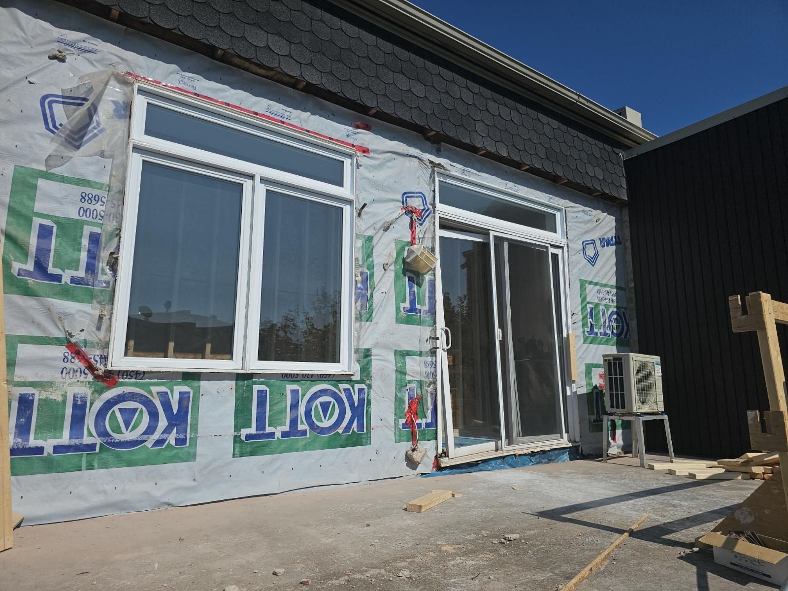 Extérieur d'une maison en construction, avec des fenêtres et des portes à cadre blanc sur un mur recouvert d'un film publicitaire.