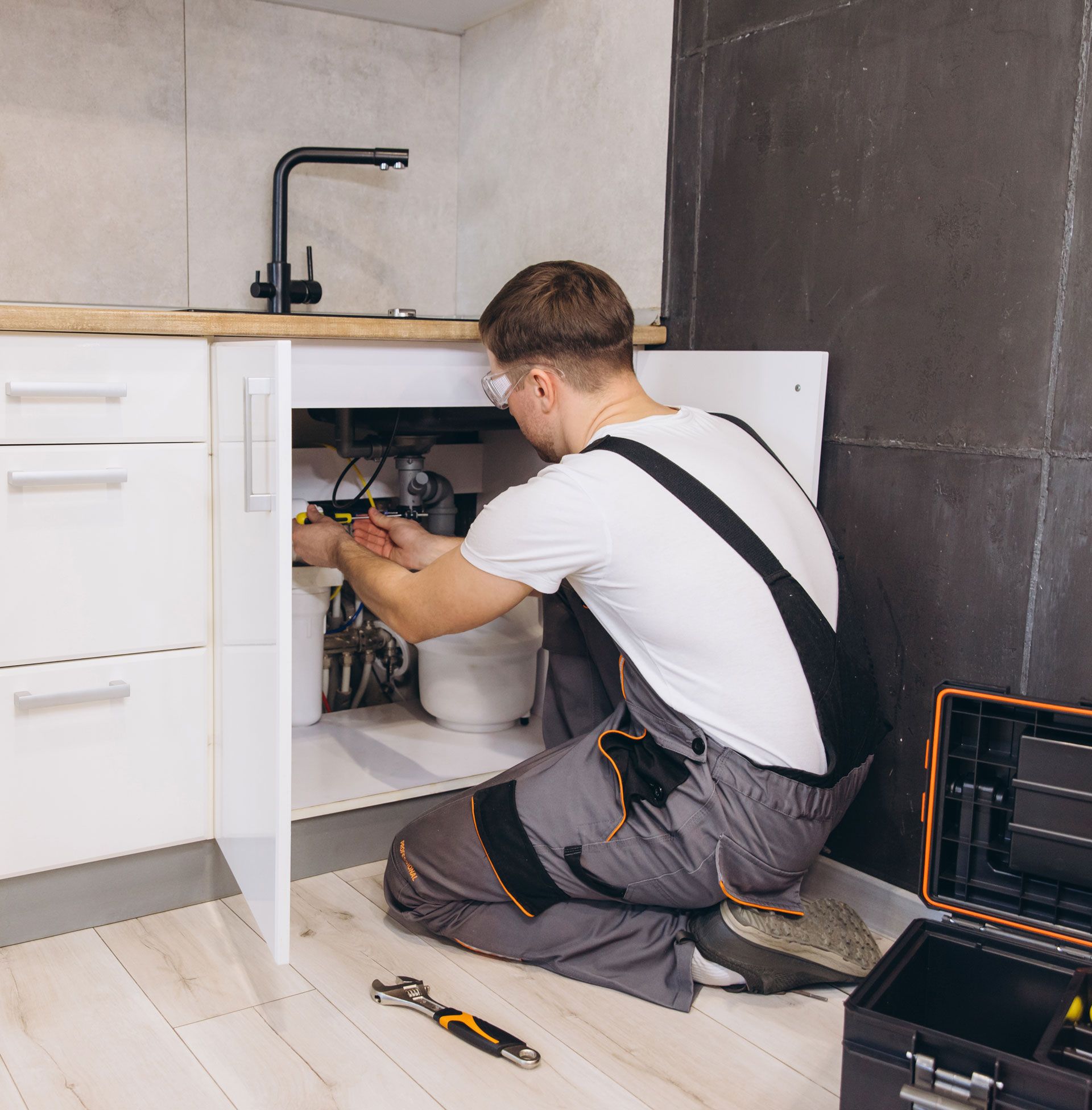 Plumber repairing pipes under a kitchen sink, with tools on the floor and cabinet doors open
