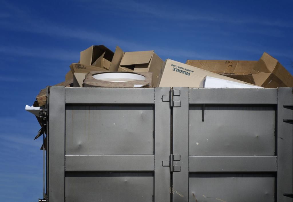 Gray dumpster overflowing with cardboard boxes under a clear blue sky