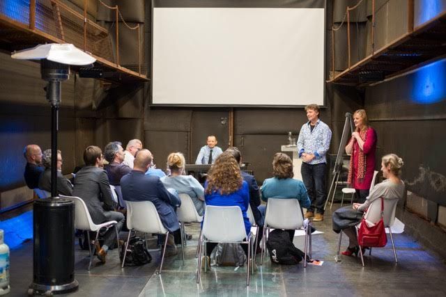 Het publiek zit en luistert naar twee presentatoren die voor een leeg scherm staan in een ruimte met industriële details.