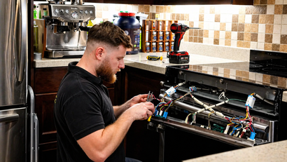 Barista working on an espresso machine in a kitchen, with coffee equipment and tiled backsplash.