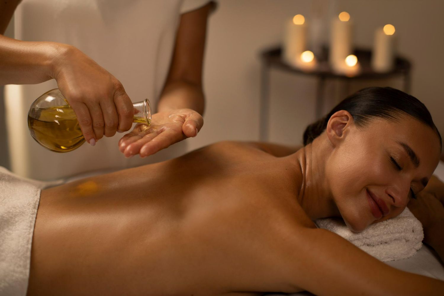 A therapist pours massage oil onto their hand before applying it to the back of a person lying on a spa massage table.